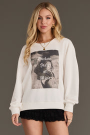 A woman with long blonde hair wears the Panache Apparel Collection’s Outlaw State of Mind Sweatshirt, a white sweatshirt featuring a black and white graphic and bold lettering, paired with black lace shorts, standing against a plain background.
