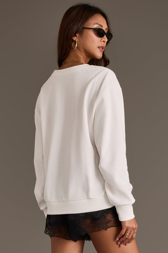 A woman wearing a Cowboy Hat Graphic Sweatshirt from Panache Apparel Collection, sunglasses, gold hoop earrings, and black lace shorts stands facing away, looking over her shoulder against a neutral background.