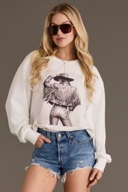 A woman wearing sunglasses, distressed denim shorts, and the Panache Apparel Collection Fringe Cowgirl Sweatshirt, featuring a graphic of a person in a fringed jacket and cowboy hat, stands against a plain background.