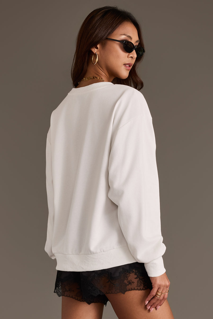 A woman wearing sunglasses and the “Better Late Than Ugly Sweatshirt” by Panache Apparel Collection, paired with black lace shorts, poses with her back to the camera against a neutral background.