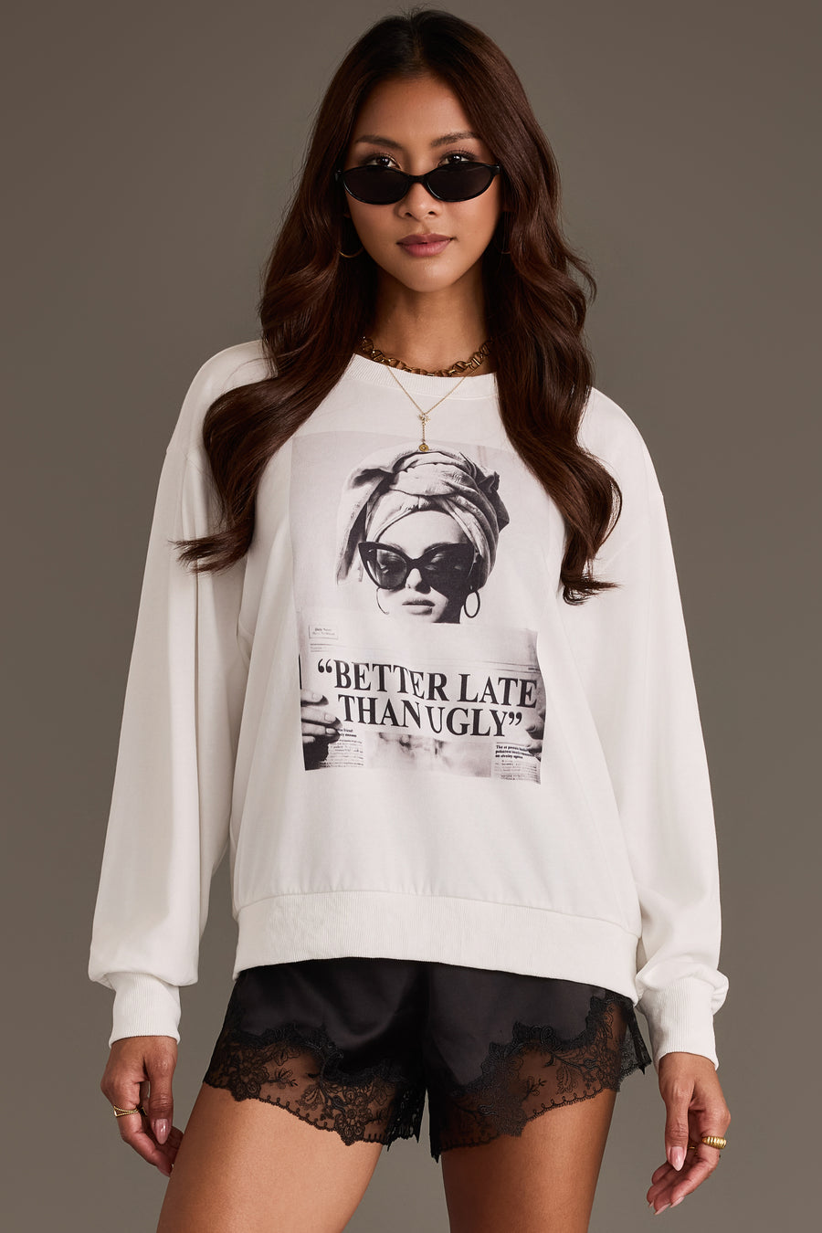 A woman wears the Panache Apparel Collection “Better Late Than Ugly” Sweatshirt, featuring a graphic print of a woman and bold text, paired with black lace shorts and sunglasses, as she stands against a plain background.
