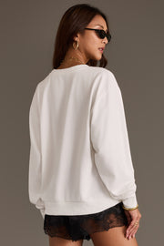 A woman stands facing away, looking over her shoulder against a plain background, wearing the Panache Apparel Collection “I Don’t Care Sweatshirt,” paired with sunglasses, gold jewelry, and black lace shorts.