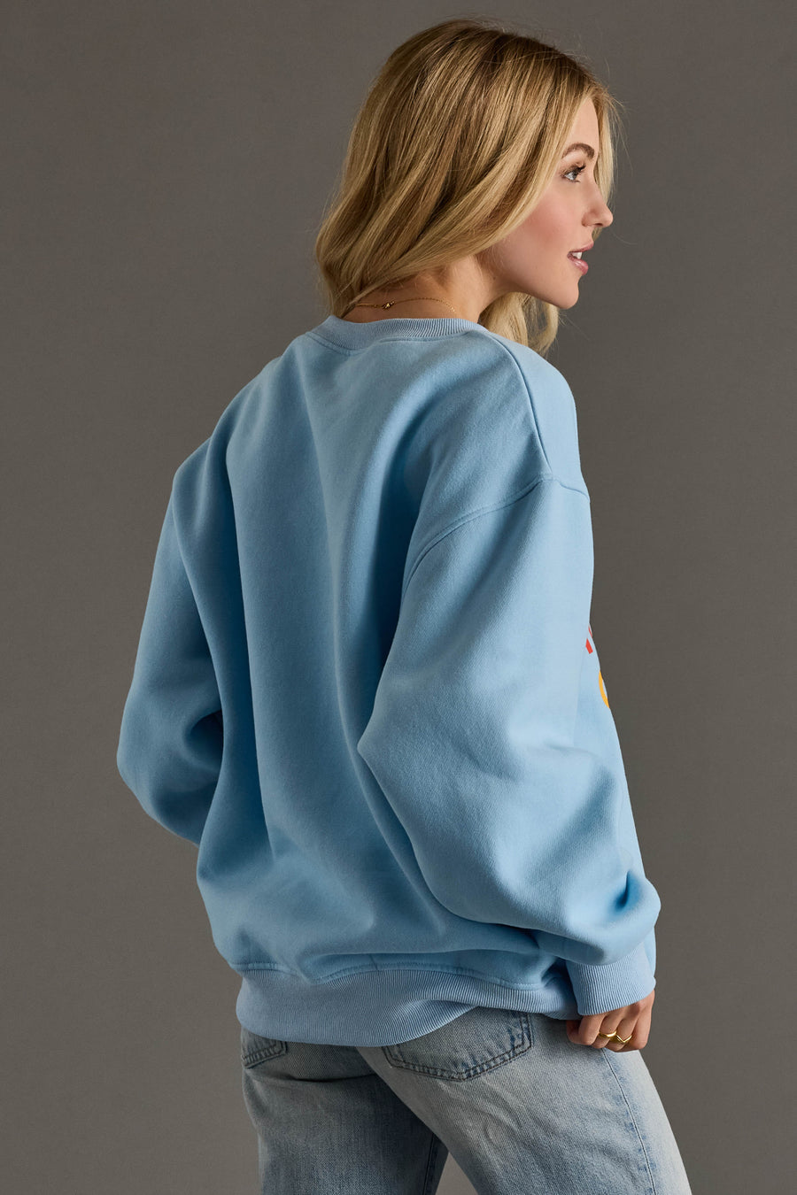 A person with long blonde hair wears the Panache Apparel Collection Light Blue Drink On A Boat Sweatshirt and light blue jeans, standing with their back to the camera and turning slightly against a plain gray background.