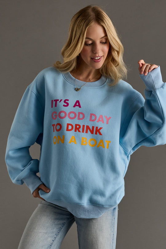 A woman with blonde hair and light jeans models the Panache Apparel Collection Light Blue Drink On A Boat Sweatshirt, featuring colorful "ITS A GOOD DAY TO DRINK ON A BOAT" text, against a plain gray background.