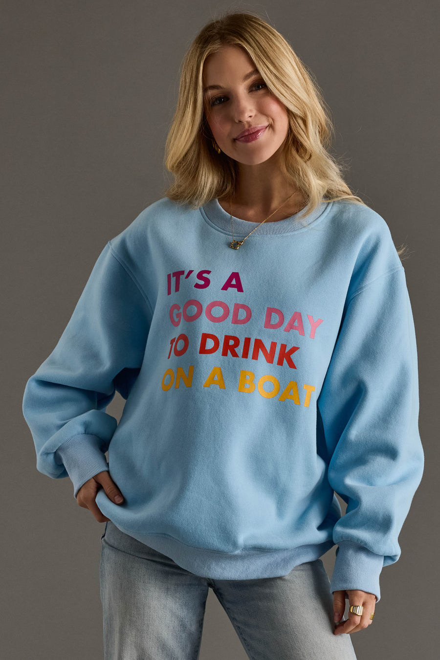 A woman smiles with one hand in her pocket, wearing the Panache Apparel Collection Light Blue Drink On A Boat Sweatshirt against a gray background. The relaxed fit top features “ITS A GOOD DAY TO DRINK ON A BOAT” in colorful letters.