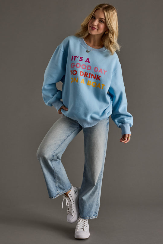 A woman smiles, one foot lifted, in a Panache Apparel Collection Light Blue Drink On A Boat Sweatshirt with "ITS A GOOD DAY TO DRINK ON A BOAT," paired with light blue jeans and white sneakers against a gray background.