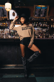A woman wearing a black hat, the Panache Apparel Collection Tan Whiskey Weather Sweater in a relaxed fit, black shorts, and cowboy boots poses in front of a bar lined with shelves of liquor bottles.