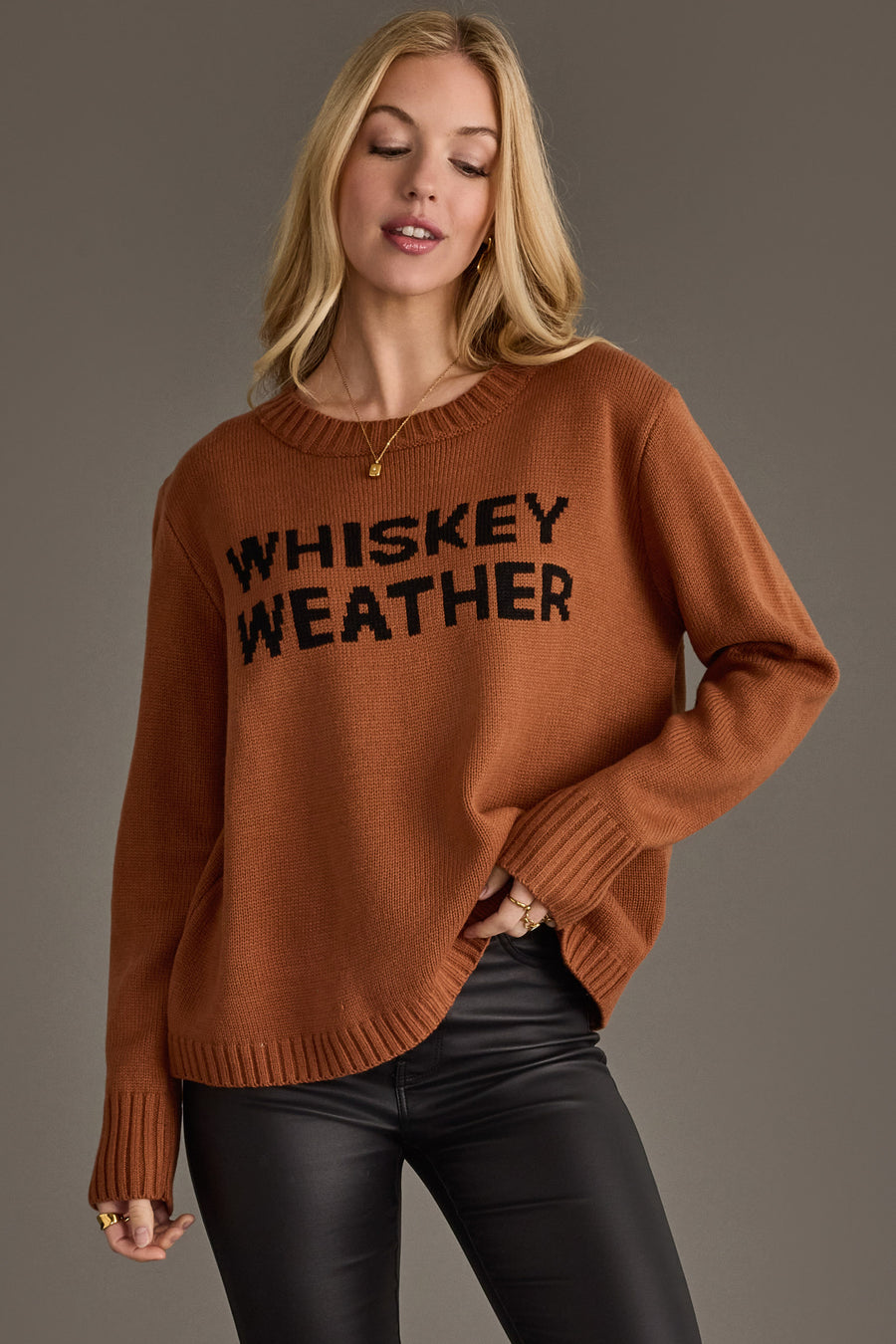 A woman with long blonde hair wears the Brown Whiskey Weather Sweater from Panache Apparel Collection, paired with black leather pants, standing against a plain gray background.