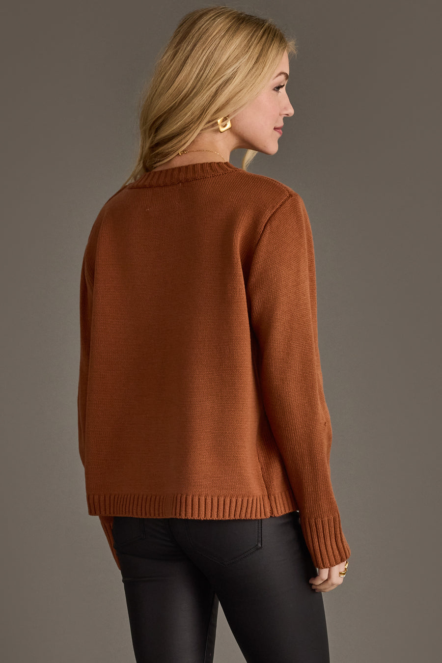 A woman with long blonde hair, wearing gold hoop earrings, a brown Whiskey Weather Sweater from the Panache Apparel Collection, and black pants stands facing away, looking over her right shoulder against a plain, neutral background.