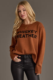 A woman with long blonde hair wears a Panache Apparel Collection Brown Whiskey Weather Sweater and black leather pants, standing against a plain background.