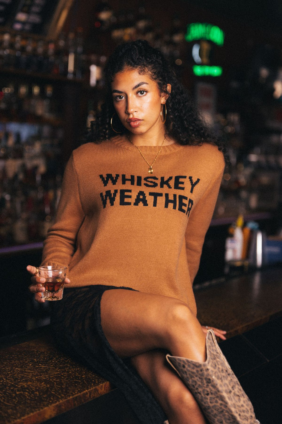 A woman with curly hair sits on a warmly lit bar counter, surrounded by softly glowing bottles. She is wearing the Panache Apparel Collection’s Brown Whiskey Weather Sweater, styled with a black skirt and leopard print boots, and holds a glass of whiskey in her hand.