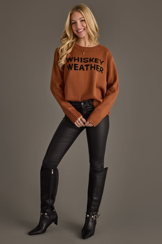 A woman stands against a gray background, smiling and wearing the Panache Apparel Collection Brown Whiskey Weather Sweater, paired with black leather pants and black knee-high boots.