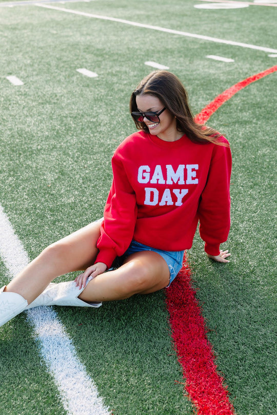 A woman wearing the Panache Apparel Collection Red Game Day Sweatshirt, denim shorts, and white shoes sits smiling on a football field near a red line, accessorized with sunglasses.
