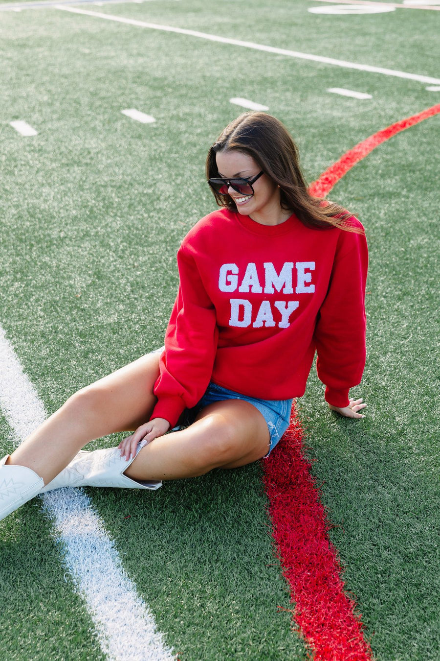 A woman wearing the Panache Apparel Collection Red Game Day Sweatshirt, denim shorts, and white shoes sits smiling on a football field near a red line, accessorized with sunglasses.