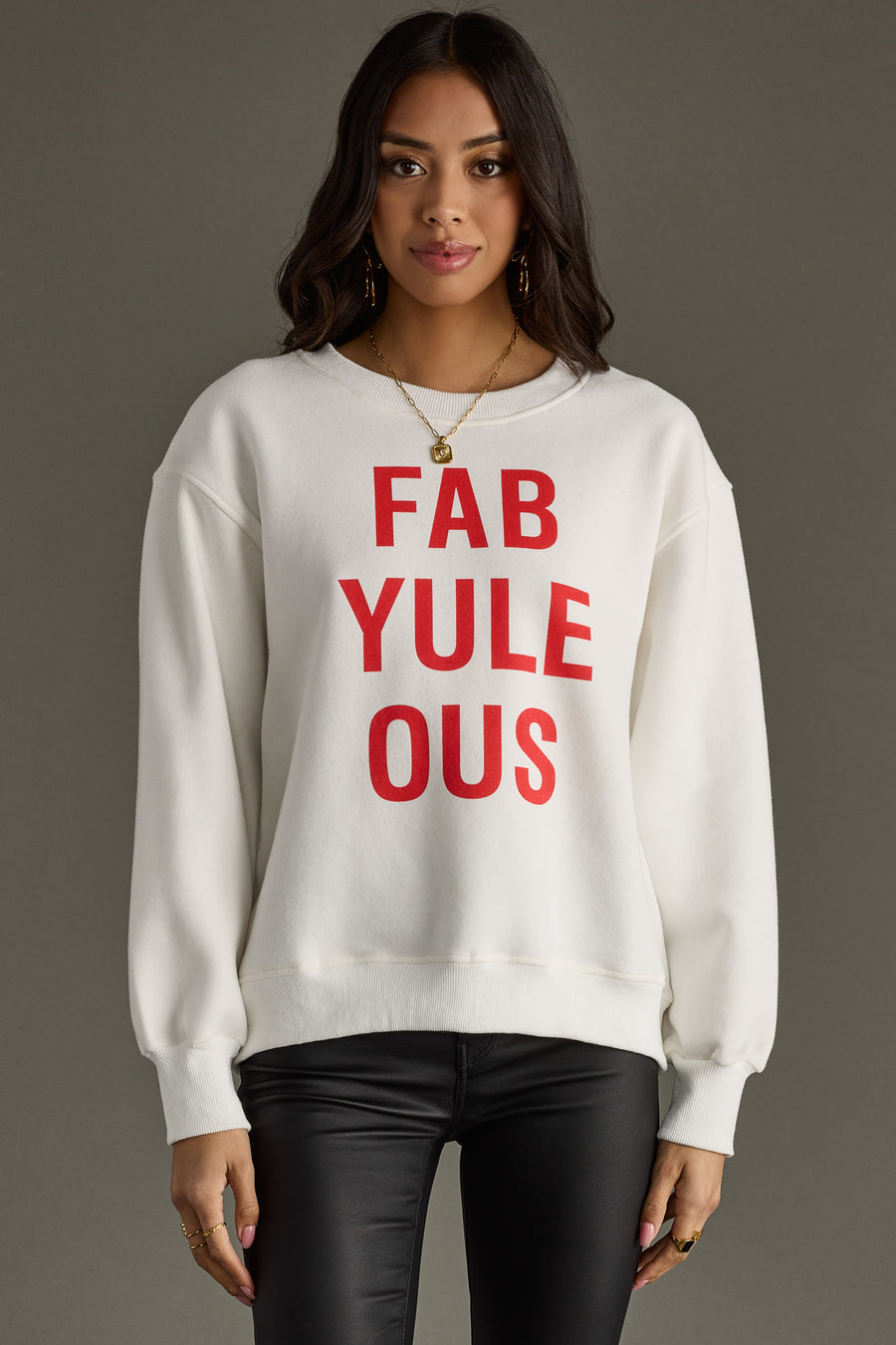 A woman in a women’s white Panache Apparel Collection FabYULEous Sweatshirt with bold red lettering stands against a gray background, wearing black pants, gold hoop earrings, and has long dark hair.