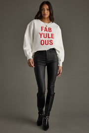 A woman with wavy brown hair and a slight smile poses against a gray background wearing the Panache Apparel Collection FabYULEous Sweatshirt in white, paired with black leather pants and knee-high boots.