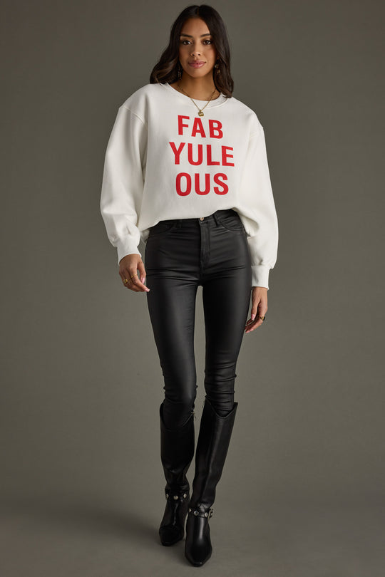 A woman with wavy brown hair and a slight smile poses against a gray background wearing the Panache Apparel Collection FabYULEous Sweatshirt in white, paired with black leather pants and knee-high boots.
