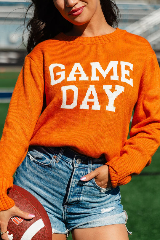 A woman stands on a sports field holding a football, dressed in the Panache Apparel Collection Orange Game Day Sweater with denim shorts for a relaxed, vibrant look.