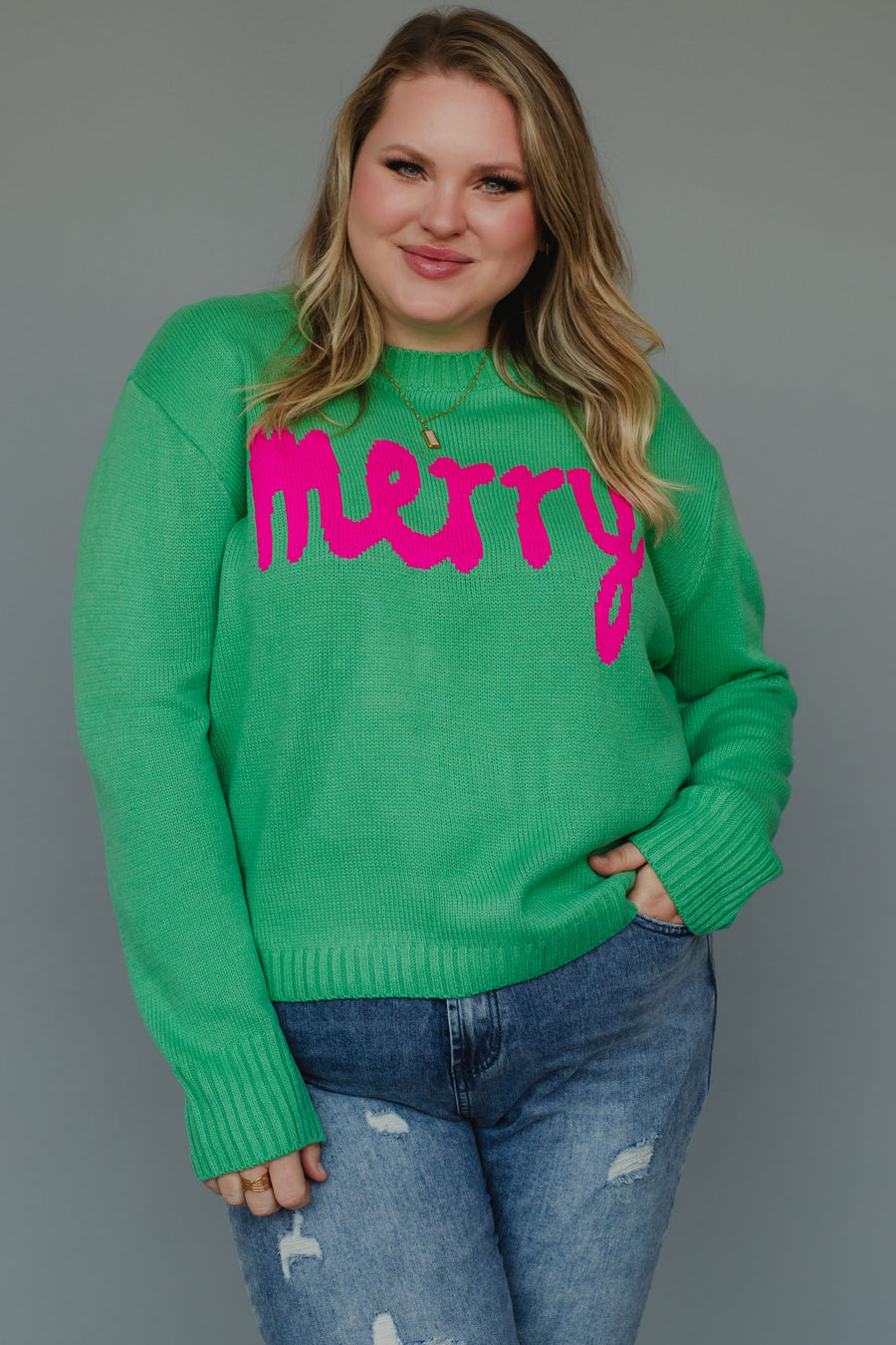 A woman wears a Mint Merry Sweater, a relaxed fit, mint green knit sweater with the word merry in bright pink letters across the chest.