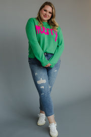 A person wears a Mint Merry Sweater in mint green knit with the word merry in pink lettering, paired with distressed blue jeans and white sneakers.