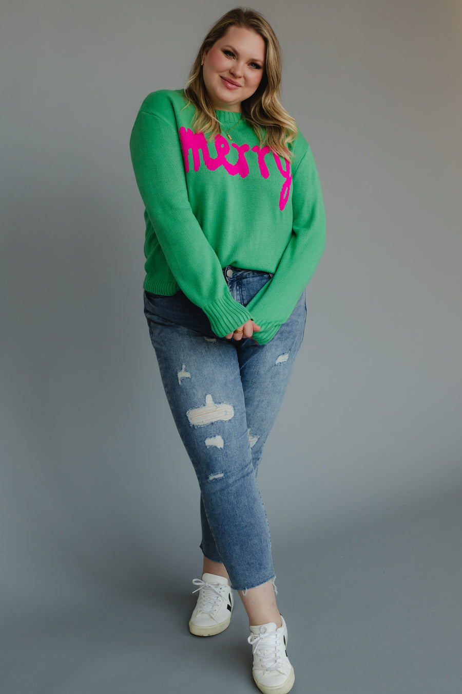 A person wears a Mint Merry Sweater in mint green knit with the word merry in pink lettering, paired with distressed blue jeans and white sneakers.