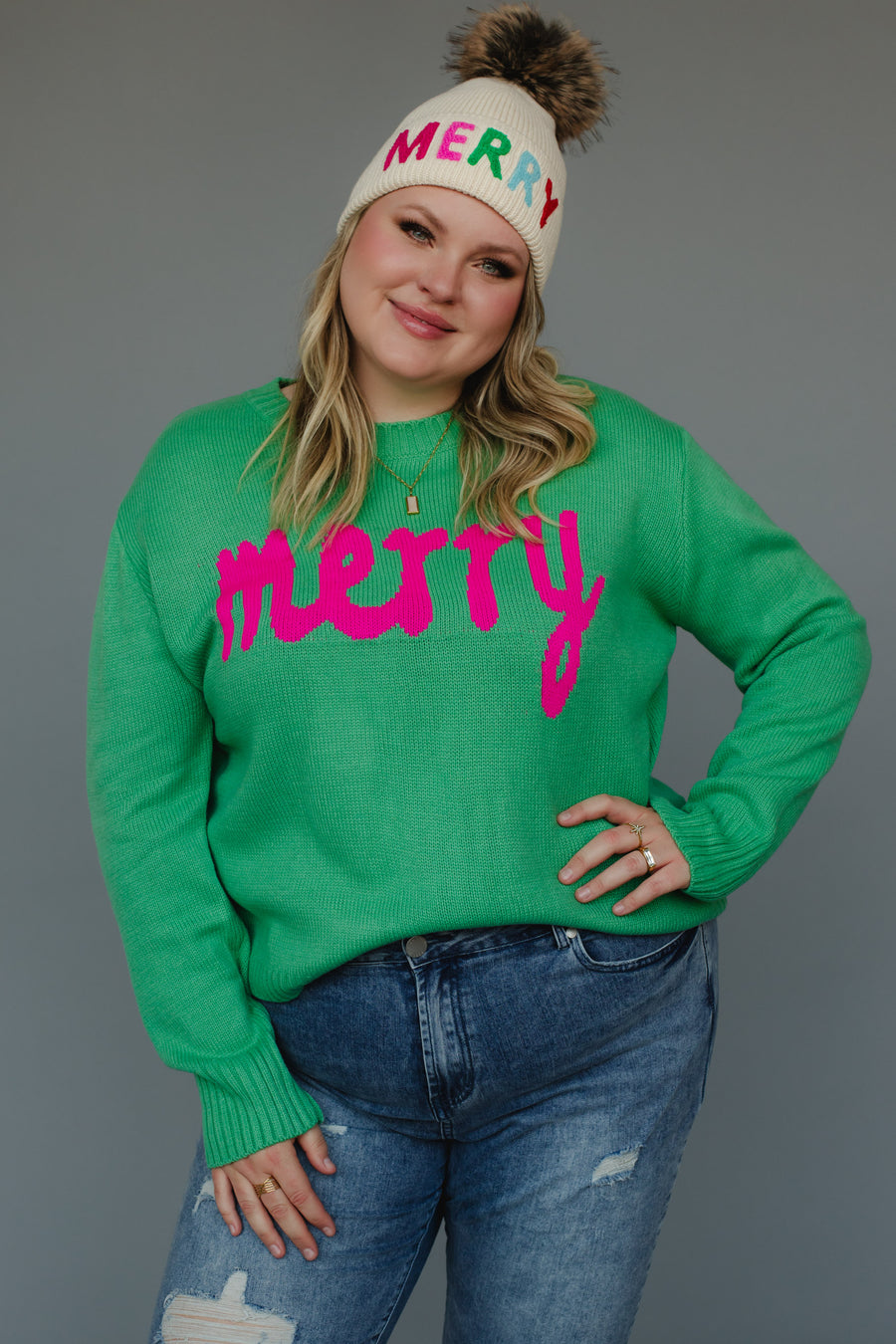 A person wears a Mint Merry Sweater in mint green with the word merry in pink, paired with a cream-colored knit beanie that has MERRY in colorful letters and a faux-fur pom-pom.