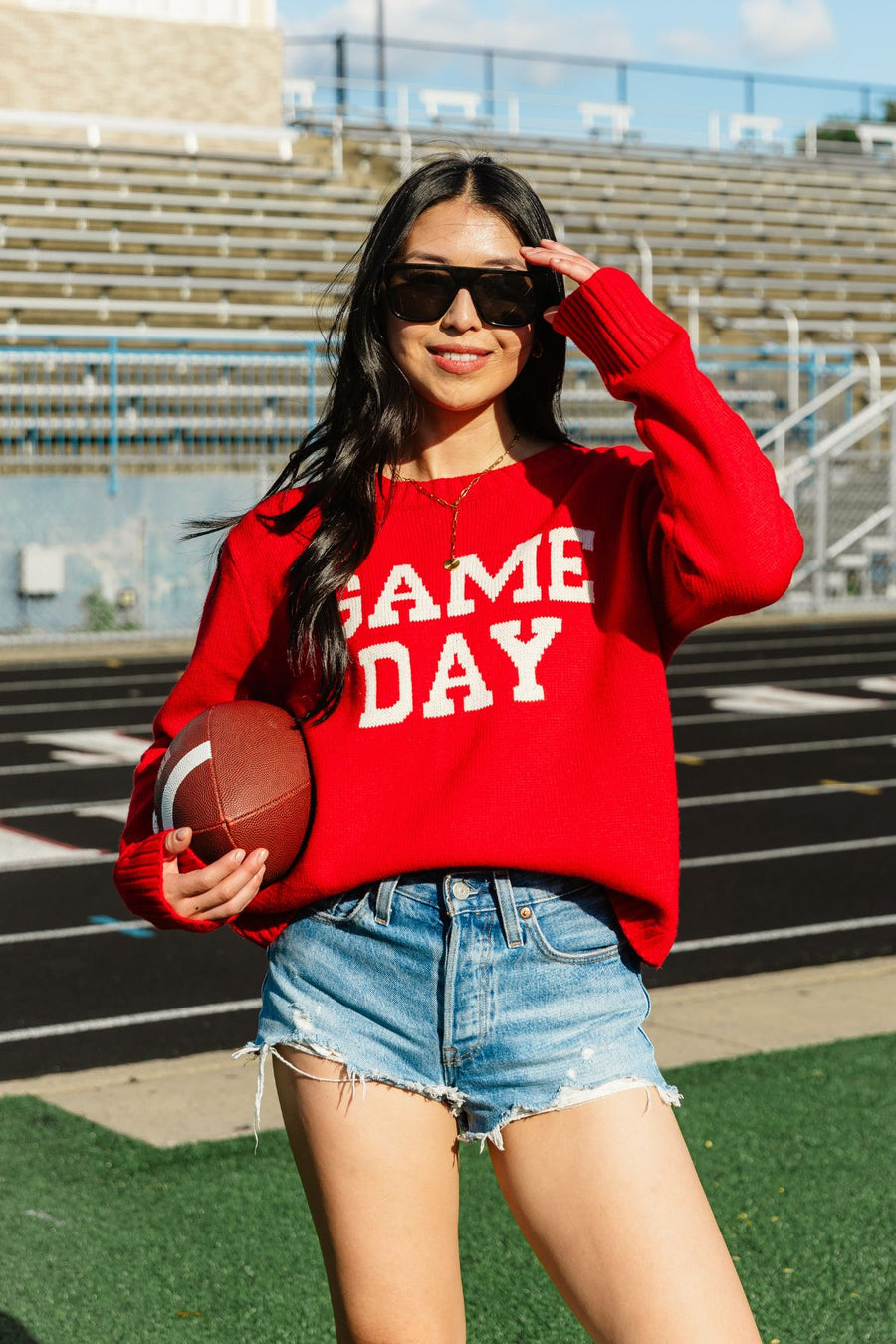 A woman wearing sunglasses and a relaxed fit Red Game Day Sweater by Panache Apparel Collection holds a football while standing on a track field with bleachers in the background, smiling at the camera.
