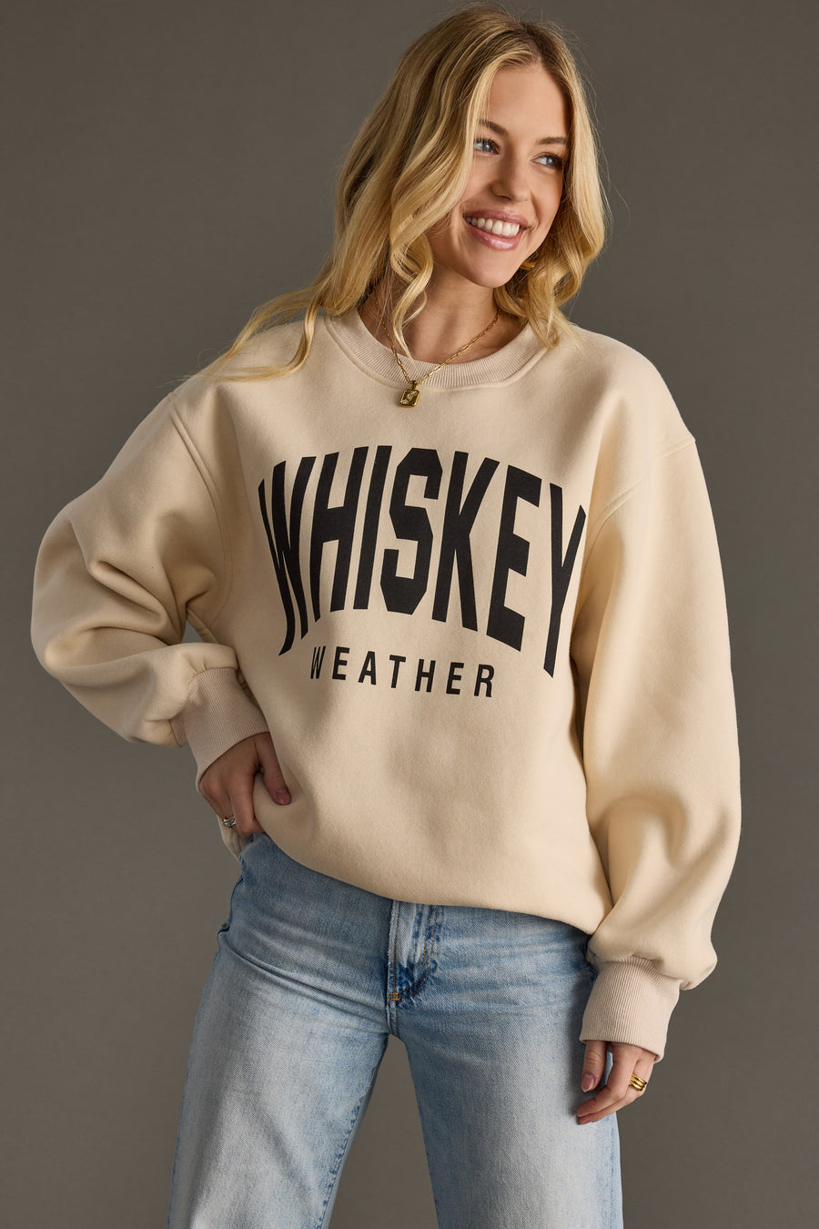 A smiling woman with long blonde hair wears the Panache Apparel Collection Whiskey Weather Sweatshirt in tan, paired with light blue jeans, standing against a plain gray background.