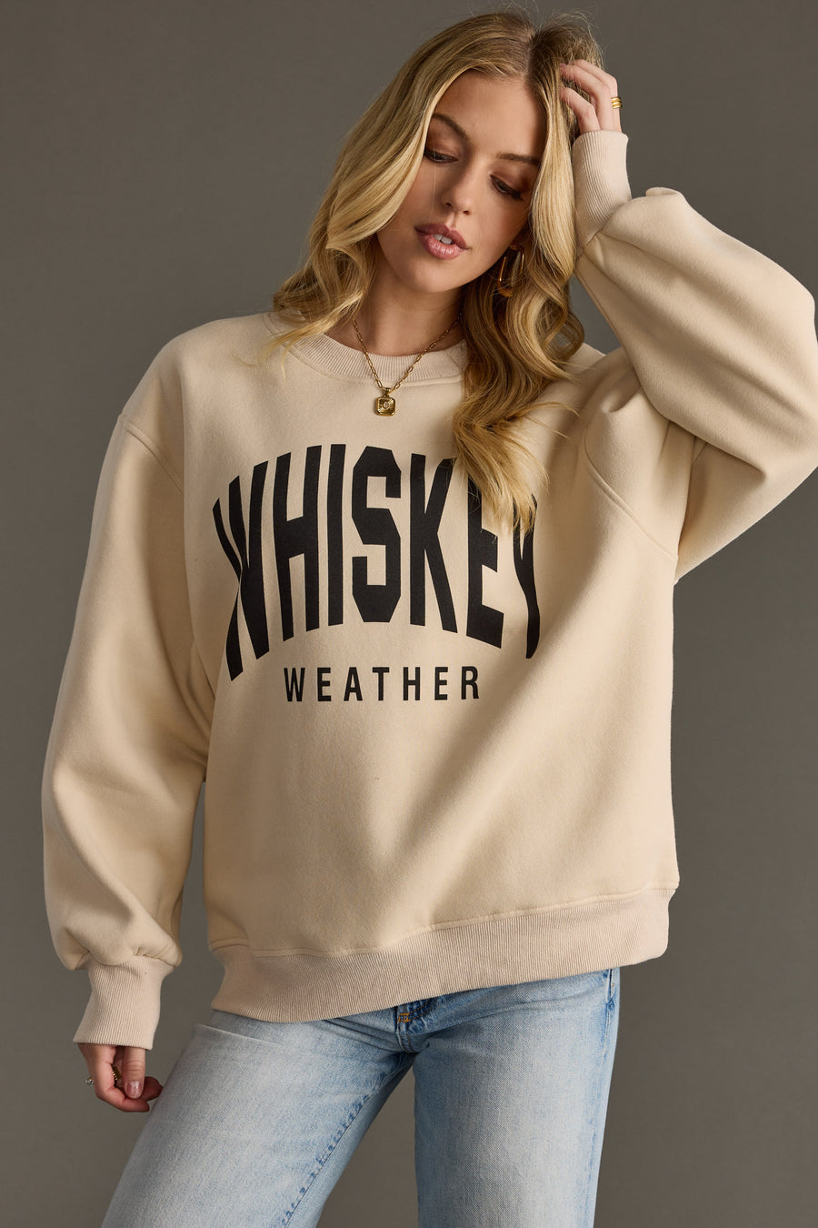 A woman in the Panache Apparel Collection Whiskey Weather Sweatshirt - Tan, featuring bold black lettering, pairs it with light blue jeans against a plain gray background.