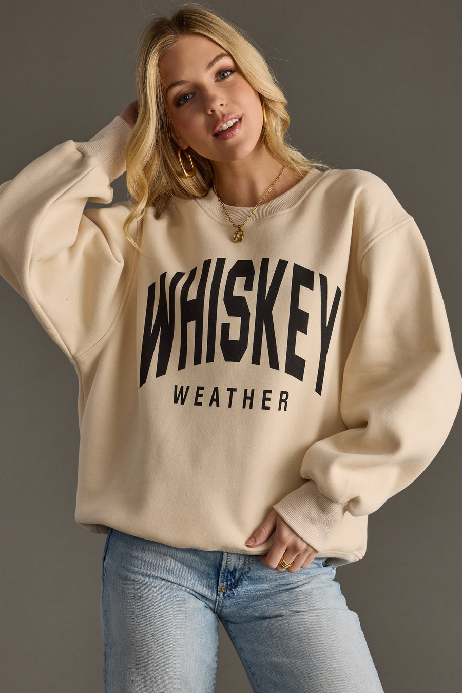 A blonde woman wears the Panache Apparel Collections Whiskey Weather Sweatshirt in tan with light blue jeans, standing against a gray background.