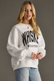 A woman with long blonde hair models the oversized Whiskey Weather Sweatshirt - White by Panache Apparel Collection, paired with light blue jeans against a plain gray background.