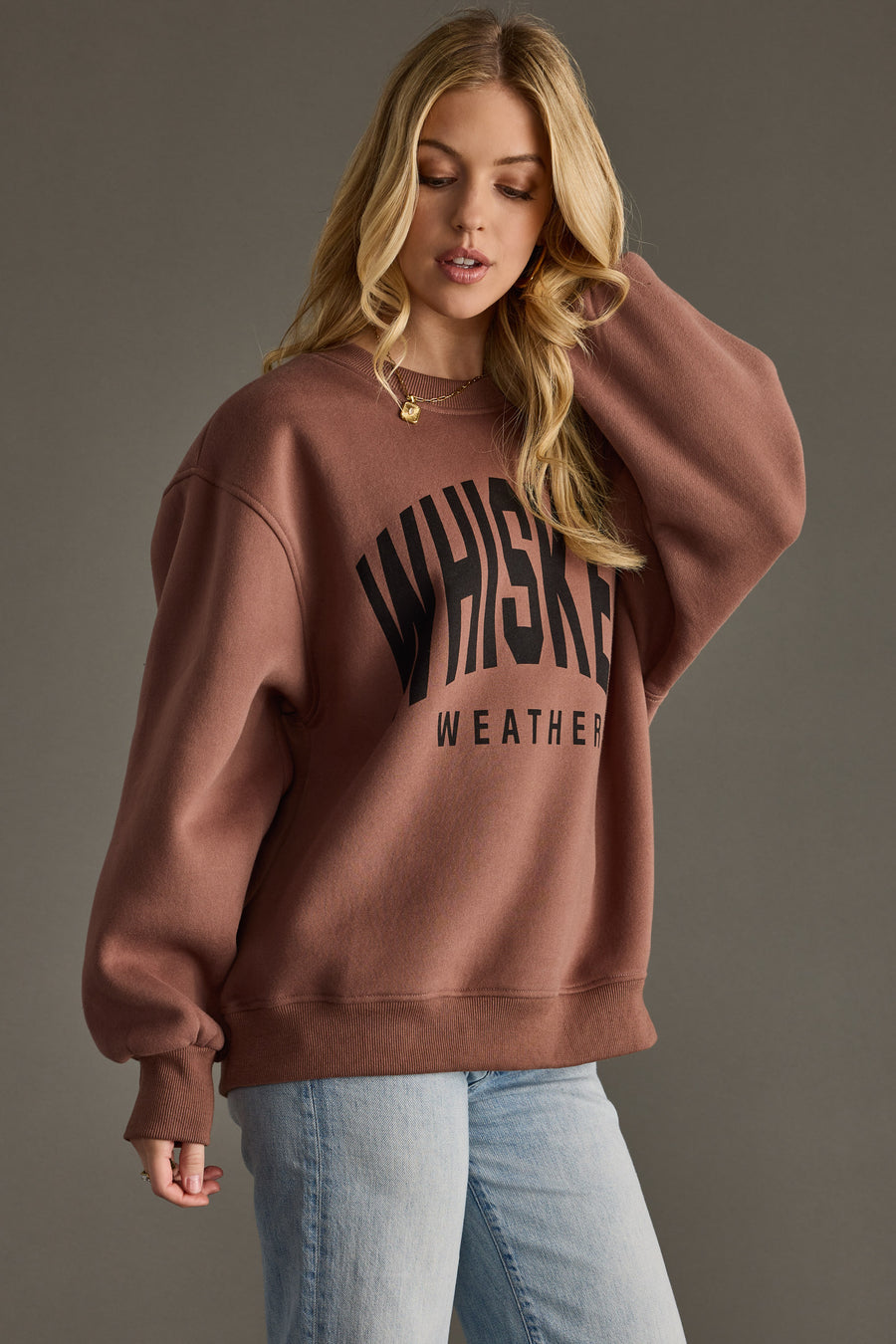 A woman with long blonde hair wears the Panache Apparel Collection Whiskey Weather Sweatshirt - Brown and light blue jeans, looking down and touching her hair with one hand against a plain background.