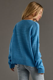 A woman with long blond hair wears the Panache Apparel Collection’s Blue Lake Life Lightweight Sweater and light-wash jeans, standing with her back turned and head slightly to the side against a plain gray background.