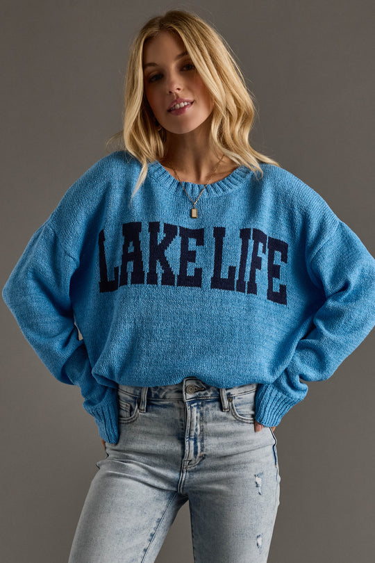 A person wears the Blue Lake Life Lightweight Sweater by Panache Apparel Collection—a blue knit, navy crew neck with bold lettering—paired with light-wash jeans, standing against a plain gray background.