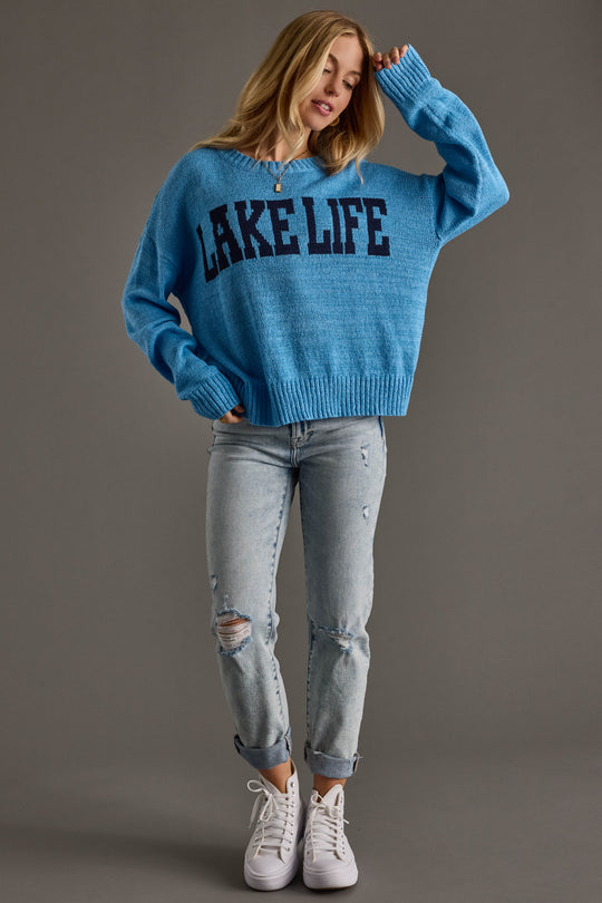 A woman with long blonde hair stands against a gray background wearing a Blue Lake Life Lightweight Sweater by Panache Apparel Collection, light ripped jeans, and white sneakers, holding one sleeve up near her face.