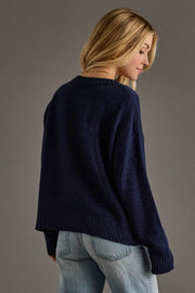 A woman with long blonde hair, wearing the Panache Apparel Collection’s Drink On A Boat Lightweight Sweater in navy blue and light blue jeans, stands slightly turned to the side against a plain gray background.
