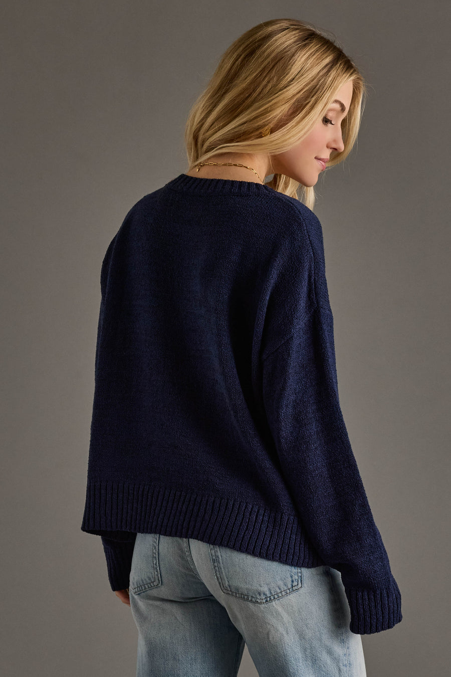 A woman with long blonde hair, wearing the Panache Apparel Collection’s Drink On A Boat Lightweight Sweater in navy blue and light blue jeans, stands slightly turned to the side against a plain gray background.