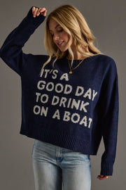 A woman in the Panache Apparel Collection's Drink On A Boat Lightweight Sweater—navy blue with "ITS A GOOD DAY TO DRINK ON A BOAT" in white—light blue jeans, and a gold necklace smiles, raising one arm against a plain background.