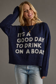 A smiling woman with long blonde hair models the Panache Apparel Collection Drink On A Boat Lightweight Sweater with "ITS A GOOD DAY TO DRINK ON A BOAT" text, paired with light blue jeans, against a plain gray background.