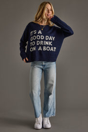 A woman models Panache Apparel Collection’s Drink On A Boat Lightweight Sweater in navy blue, styled with light blue jeans and white sneakers against a plain gray background.