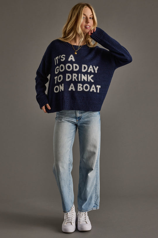 A woman models Panache Apparel Collection’s Drink On A Boat Lightweight Sweater in navy blue, styled with light blue jeans and white sneakers against a plain gray background.