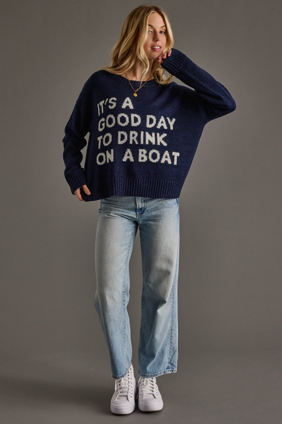 A woman models Panache Apparel Collection’s Drink On A Boat Lightweight Sweater in navy blue, styled with light blue jeans and white sneakers against a plain gray background.