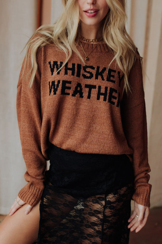 A woman wears the Brown Whiskey Weather Lightweight Sweater from Panache Apparel Collection with a black lace skirt, standing indoors before light-colored curtains.