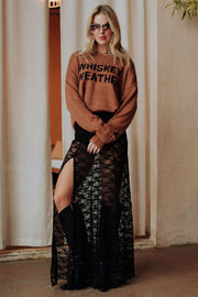 A woman in sunglasses, a Panache Apparel Collection Brown Whiskey Weather Lightweight Sweater, and a black lace skirt with high slits stands indoors on a concrete floor before cream-colored curtains.