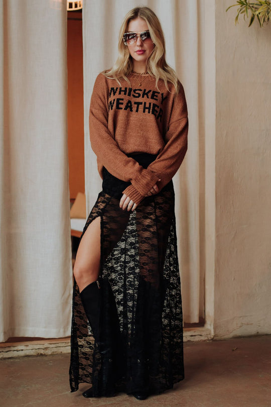 A woman in sunglasses, a Panache Apparel Collection Brown Whiskey Weather Lightweight Sweater, and a black lace skirt with high slits stands indoors on a concrete floor before cream-colored curtains.