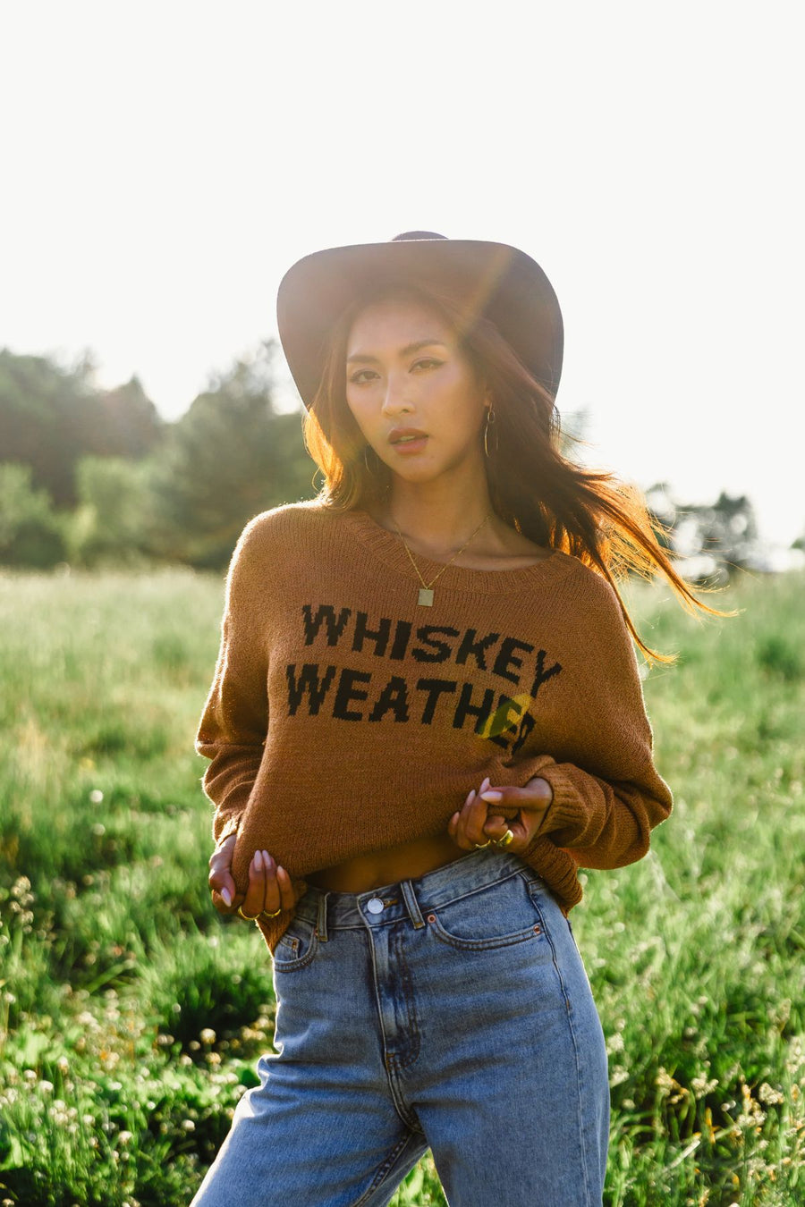 A woman wearing a hat and the Panache Apparel Collection Brown Whiskey Weather Lightweight Sweater stands in a sunny field of green grass, looking at the camera with one hand on her waist and the other holding her sweater.
