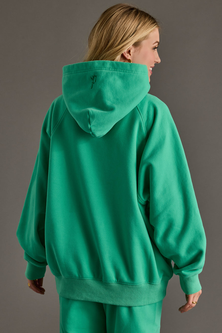A woman with long blonde hair stands with her back to the camera, wearing the Green Panache Hooded Sweatshirt from Panache Apparel Collection. The cozy fleece hoodie and matching pants stand out against a plain gray background.