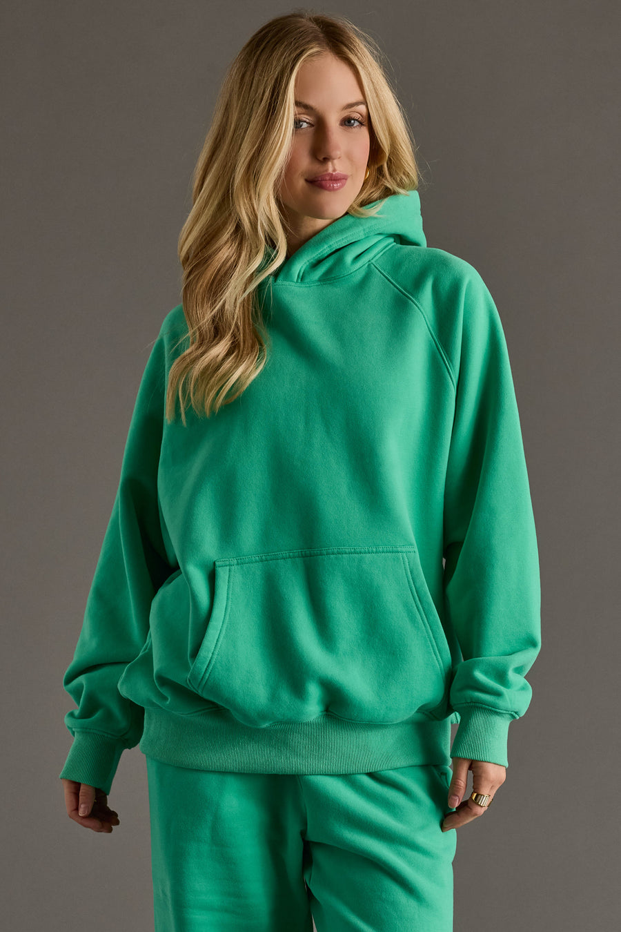 A woman with long blonde hair wears the oversized Green Panache Hooded Sweatshirt from Panache Apparel Collection, paired with matching green pants, standing against a plain gray background.