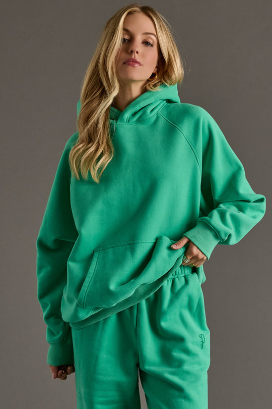 A woman with long blonde hair wears the Green Panache Hooded Sweatshirt by Panache Apparel Collection and matching sweatpants, standing against a plain gray background.