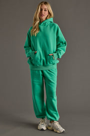 A person with long blond hair stands against a gray background wearing the Panache Apparel Collection's Green Panache Hooded Sweatshirt and matching sweatpants, paired with white sneakers and hands in the hoodie’s front pocket.